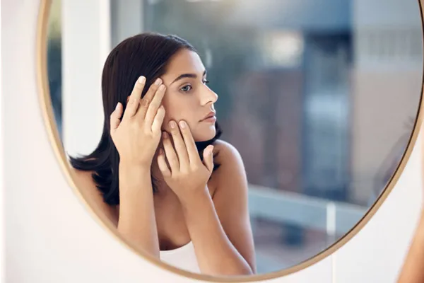 Dziewczyna o brązowych włosach przygląda się swojej skórze pod oczami przed lustrem 